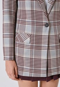 Geruit blazer in grijs, bordeauxrood en lichtblauw. Heeft twee voorzakken en een enkele knoopsluiting. De stof heeft een gemêleerd uiterlijk.
