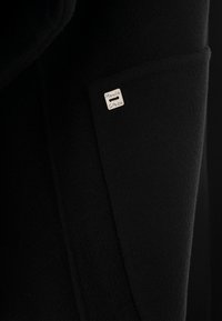Cappotto nero realizzato in un tessuto liscio e strutturato, con un'etichetta logo argentata sulla tasca. Design semplice con una silhouette raffinata.