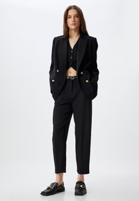 Blazer nero su misura con doppi bottoni dorati, top corto nero con bottoni, pantaloni neri affusolati e mocassini neri con accenti dorati.