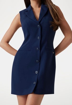 Mouwloze marineblauwe jurk met knopen aan de voorkant, reverskraag, getailleerde taille en zijzakken gedragen door een persoon met lang bruin haar.