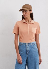 Perzikkleurig polo-shirt met een oplopende kraag, knoopjes aan de voorkant tot halverwege en een zak; gecombineerd met lichtblauwe wijde jeans. Een bruine suède pet maakt de outfit compleet.