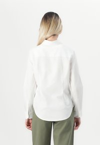 Person with long blonde hair wearing a white long-sleeve shirt and olive green pants, viewed from the back against a white background.