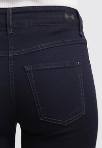Mørkeblå denimjeans med et tettsittende design. Funksjoner inkluderer baklommer og kontrasterende sømmer. Merking synlig på linningen.
