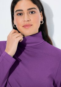 Lila Langarmoberteil mit hohem Kragen, aus glattem Stoff. Verfügt über figurbetonte Ärmeln und eine genähte Dekoration am Kragen.