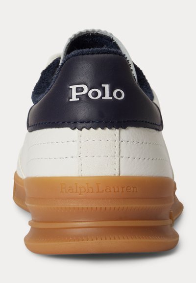 Οπίσθια όψη λευκού αθλητικού παπουτσιού με μπλε ταμπελάκι στη φτέρνα που γράφει «Polo» και ταμπά σόλα με ανάγλυφο το όνομα «Ralph Lauren».