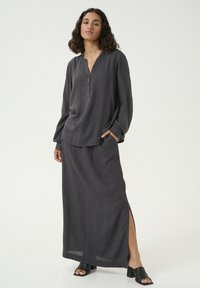 Top in lino grigio con bottoni e maniche lunghe, abbinato a una gonna maxi coordinata con spacchi laterali. Sandali neri con tacco completano il look.