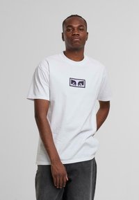 Hvid bomulds t-shirt med rund hals og korte ærmer, udsmykket med et sort og lilla grafisk logo på brystet. Bæres med grå bukser.