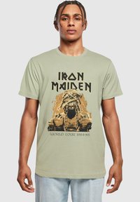 Lys grøn t-shirt i bomuld med et grafisk motiv af en indhyllet figur med kæder, samt "IRON MAIDEN WORLD TOUR 1984-85" i fed sort skrift.