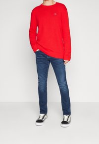 Roter Strickpullover mit gerippten Bündchen und Saum, kombiniert mit blauen Slim-Fit-Jeans und schwarz-weißen Sneakers. Einfaches Logo auf dem Pullover.