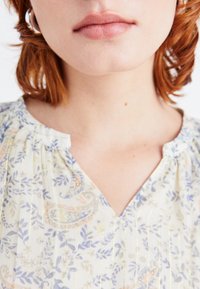 Blusa floreale in tessuto leggero, con scollatura a V e pieghe. I colori includono motivi pastello di foglie e paisley su uno sfondo crema.