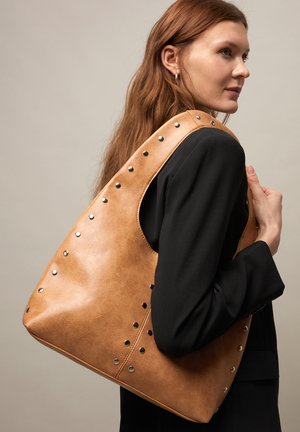 Young woman with long reddish hair wearing black blazer carries large tan leather shoulder bag with metal studs.