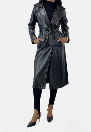 Femme portant un long trench-coat noir en cuir synthétique avec une ceinture nouée, leggings noirs et chaussures à talons pointus noires, debout les mains dans les poches.
