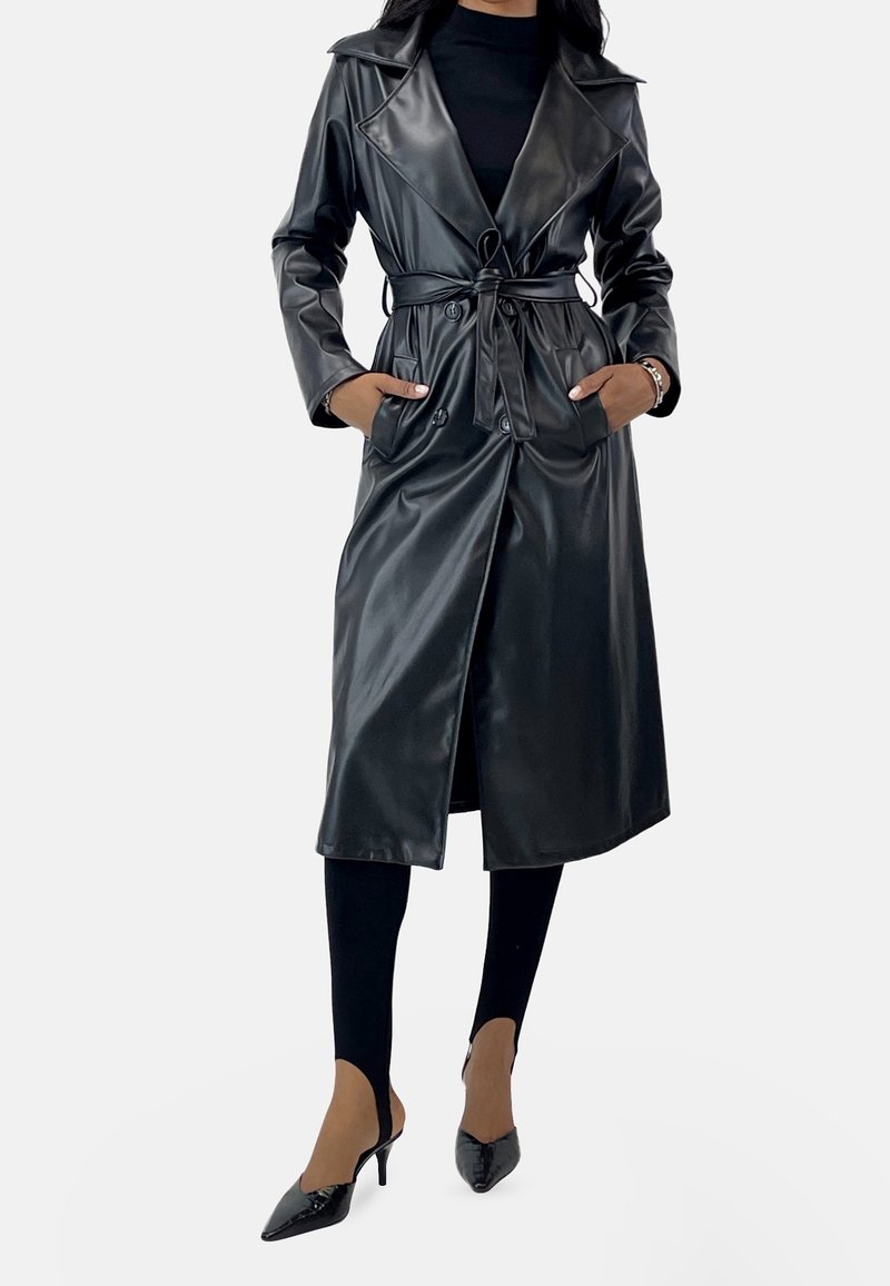 Femme portant un long trench-coat noir en cuir synthétique avec une ceinture nouée, leggings noirs et chaussures à talons pointus noires, debout les mains dans les poches.