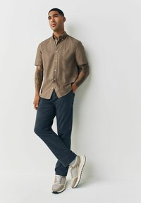 Korte mouwen bruin knoopshirt, marineblauwe broek en beige sneakers met grijze accenten. Glad materiaal met een relaxte pasvorm. Zichtbare tatoeages op de armen.