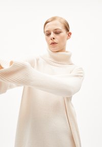 Lichtbeige coltrui met ribgebreide mouwdetails, zachte textuur en een losse pasvorm, weergegeven op een model dat zijn armen uitstrekt tegen een witte achtergrond.