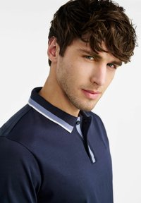 Granatowa koszulka polo z teksturowanym, białym i niebieskim kołnierzykiem w paski, krótkimi rękawami i zapięciem na dwa guziki; gładka tkanina i dopasowany krój.