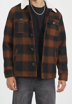 Mann trägt eine braun-schwarz karierte Knopfleistenjacke mit weißem Fleecekrause, schwarzes Hemd, silberne Kettenhalskette und graue Jeans.