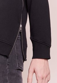 Felpa nera con zip laterale e hardware in argento, polsini a coste e texture liscia. Il design presenta una vestibilità moderna.