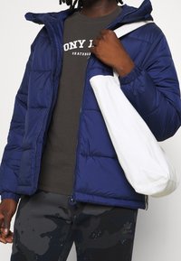 Marintröja med huva i puffer-stil, texturerad yta och elastiska ärmar; grå t-shirt under med vit text; vit tote-väskan och camouflagedbyxor.