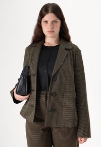 Brun og grønn mønstrete blazer med store knapper, paret med en svart topp og bukser, holder en liten svart veske.