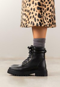 Botas de combate de cuero negro con suela de goma gruesa, diseño de cordones, detalles en hardware dorado y un calcetín texturizado gris sobre el tobillo.