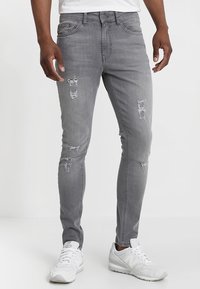 Jeans de mezclilla gris con un corte ajustado, que presentan detalles desgastados, incluyendo desgarros en la parte frontal. Combinados con zapatillas deportivas blancas.