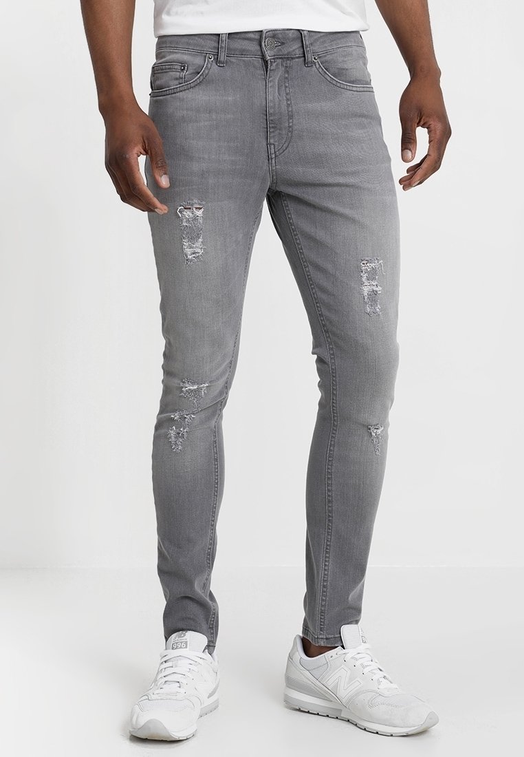 Jeans de mezclilla gris con un corte ajustado, que presentan detalles desgastados, incluyendo desgarros en la parte frontal. Combinados con zapatillas deportivas blancas.