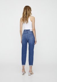 Jean en denim bleu taille haute avec revers, deux poches arrière et une étiquette sur la taille. Porté avec un débardeur blanc.