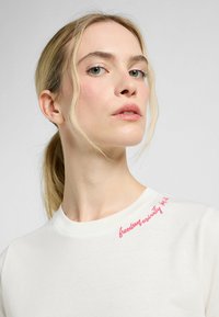 Witte katoenen t-shirt met een ronde hals en roze geborduurde tekst op de schouder. Eenvoudig ontwerp met een gladde textuur.