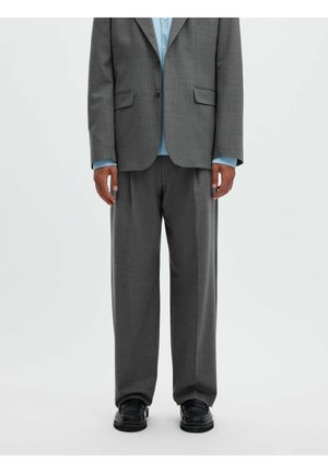 Personne portant un costume gris à fines rayures avec un pantalon assorti et une chemise bleu clair, debout devant un fond blanc uni.