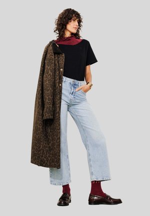 Femme aux cheveux bouclés portant un t-shirt noir, un jean large bleu clair, une écharpe bordeaux et des chaussettes, tenant un manteau en léopard brun sur son épaule.