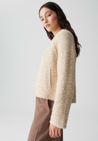 Cremefarbener, strukturierter Cardigan mit langen Ärmeln, feinen Sprenkeln und seitlichen Taschen, kombiniert mit braunen, taillierten Hosen.