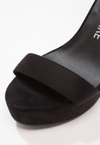 Chaussure à talon plateforme en daim noir avec une large bride sur l'avant-pied et une doublure intérieure lisse et mate. La chaussure présente un bout arrondi.