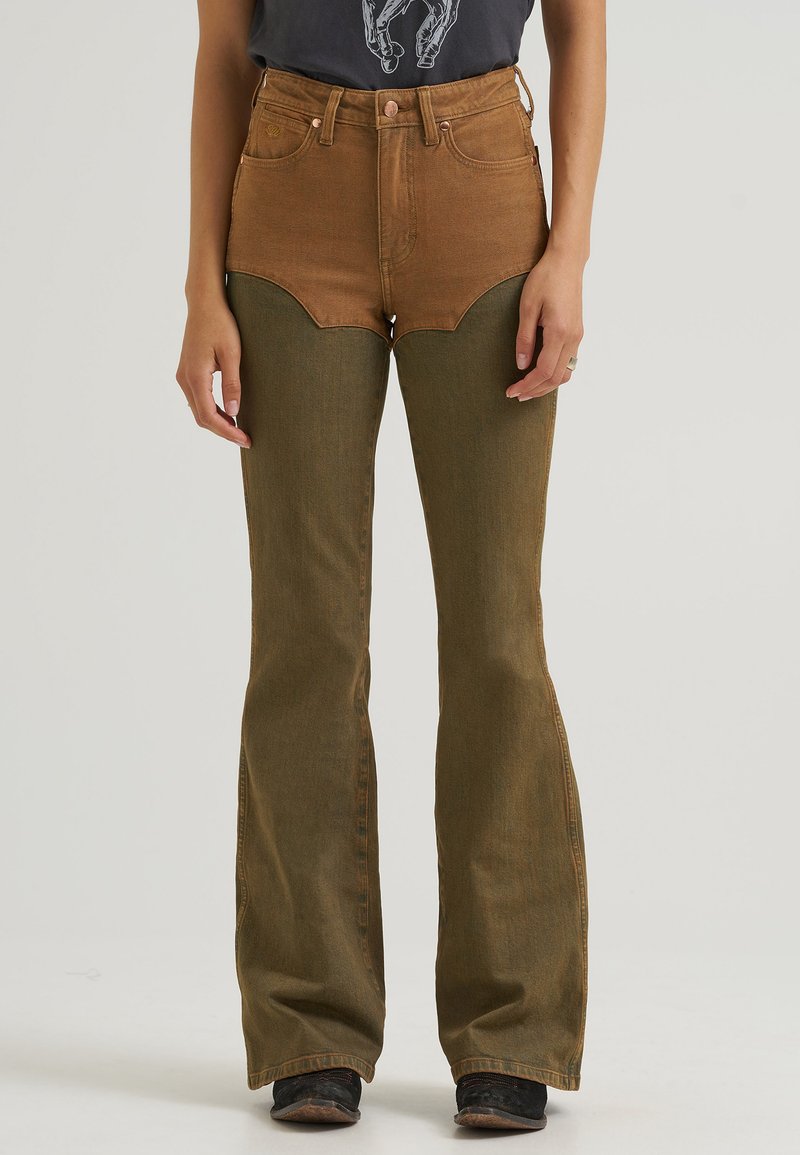 Femme portant un jean flare taille haute marron et vert olive avec poches avant, debout, les bras relâchés le long du corps.
