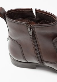 Bottine en cuir marron avec fermeture éclair latérale, couture visible et languette de tirage sur fond blanc.