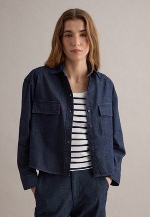 Jeune femme portant une veste en jean bleu foncé et un pantalon assorti avec un chemisier à rayures blanches et noires, posant devant un fond uni.