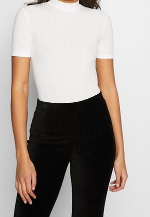 Kobieta ubrana w dopasowany biały prążkowany top z krótkim rękawem, wpuszczony w czarne prążkowane spodnie z wysokim stanem, stojąca na jednolitym tle.