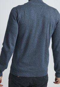 Osoba w ciemnoniebieskim swetrze z długim rękawem typu golf oraz czarnych spodniach, pokazana od tyłu na jednolitym tle.