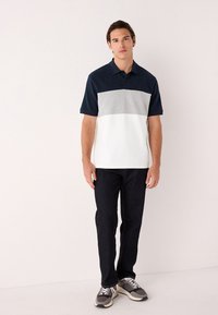 Poloshirt mit einem dunkelblauen oberen Bereich, hellgrauer Mitte und weißem unteren Abschnitt; kurze Ärmel und Standardkragen. Kombiniert mit dunkler Jeans.
