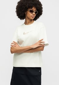 Νεαρή γυναίκα με μεγάλα σγουρά μαλλιά που φοράει μπεζ γυαλιά ηλίου, λευκό t-shirt Nike με μεταλλικό λογότυπο και μαύρη φούστα, με τα χέρια σταυρωμένα.