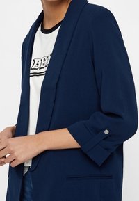 Granatowa marynarka wykonana z gładkiej tkaniny, z pojedynczym zapięciem na guzik i rękawami podwiniętymi z zatrzaskiem. Widoczna kieszeń z przodu.