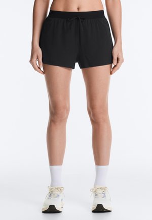 Kvinde iført sorte træningsshorts med snøre, hvide ankelsokker og hvide løbesko, stående mod en ensfarvet hvid baggrund.
