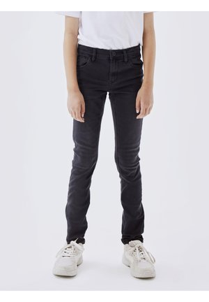 Person trägt schlanke schwarze Jeans, weißes T-Shirt und weiße Turnschuhe und steht vor einem schlichten hellen Hintergrund.