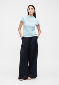 Γυναίκα που στέκεται με τα χέρια στις τσέπες, φορώντας φωτεινό μπλε κοντομάνικο τοπ, φαρδιά μαύρη παντελόνια και μπεζ σανδάλια σε λευκό φόντο.