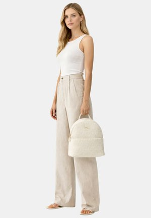 Donna bionda con top bianco senza maniche e pantaloni beige a gamba larga che tiene uno zaino crema con motivo monogramma dalla maniglia superiore.