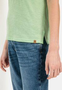 Lichtgroene kortegalm shirt met een gestructureerde afwerking, zijsplitten en een klein bruin logotag. Gemaakt in combinatie met blauwe spijkerbroek.