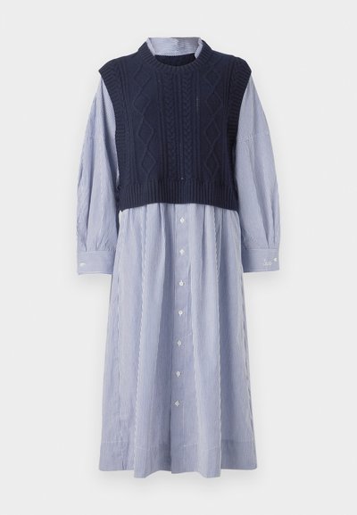 Vesta din tricot cu model cablu, de culoare bleumarin, peste o rochie cămașă cu dungi albastre și albe. Prezintă mâneci lungi și o tiv ușor evazat.