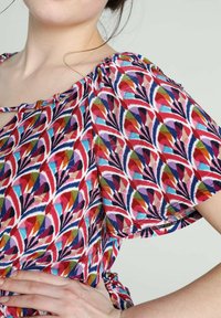 Femme portant un chemisier à manches courtes coloré avec des formes géométriques rouge, bleu, violet et vert.