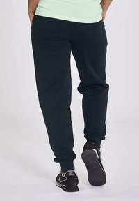 Pantalones de chándal negros con cintura elástica y puños, hechos de una tela suave. Llevados con zapatillas deportivas negras con acentos blancos.