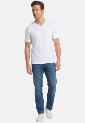 Homme marchant, portant un t-shirt blanc uni à col en V, un jean bleu et des baskets blanches, sur un fond clair uni.